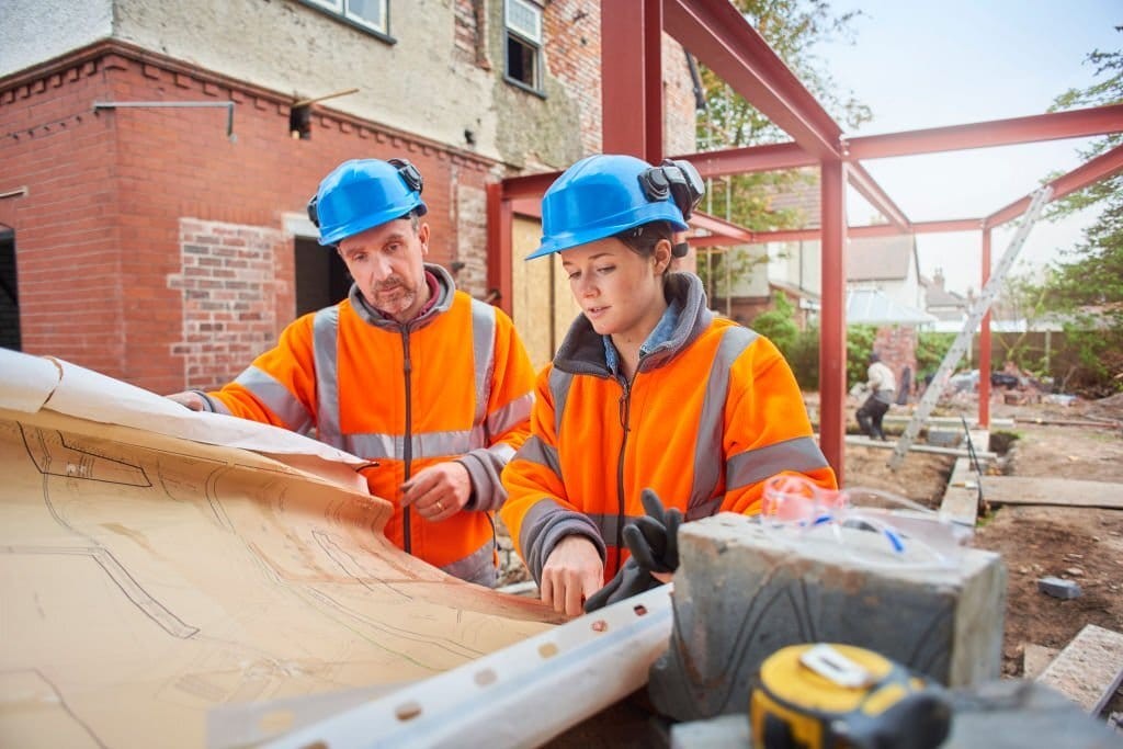 Two construction workers with safety helmets discussing blueprints at a house extension and renovation site with steel framework in the background.
