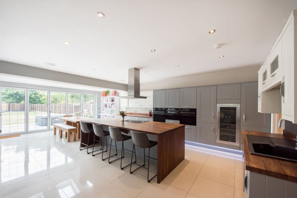 Spacious and well-lit modern kitchen extension featuring gray cabinetry, glossy porcelain floor tiles, a central island with wooden countertops and bar stools, stainless steel appliances, and sliding glass doors leading to a landscaped backyard.