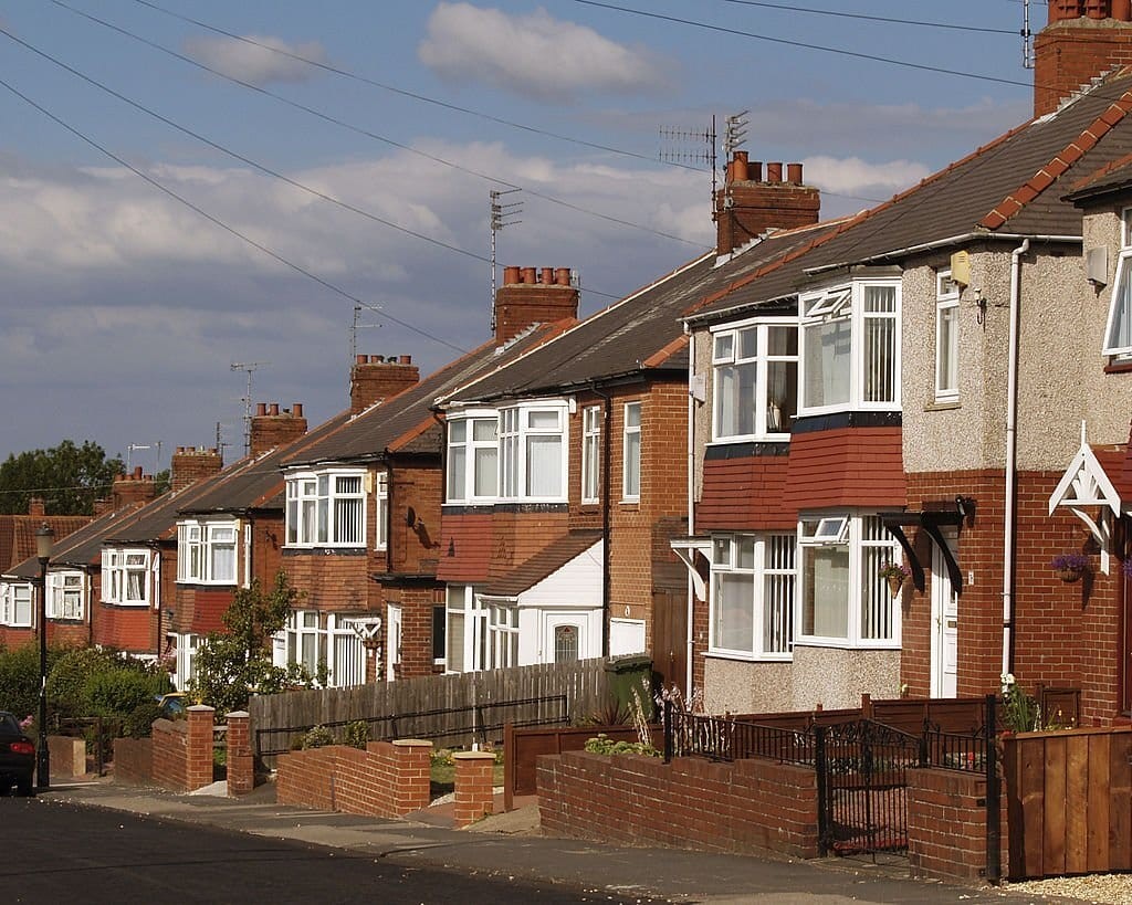 Traditional British 1930s terraced houses with distinctive red brickwork, white bay windows, and gabled roofs, on a quiet suburban street, showcasing home extensions and front garden fences.