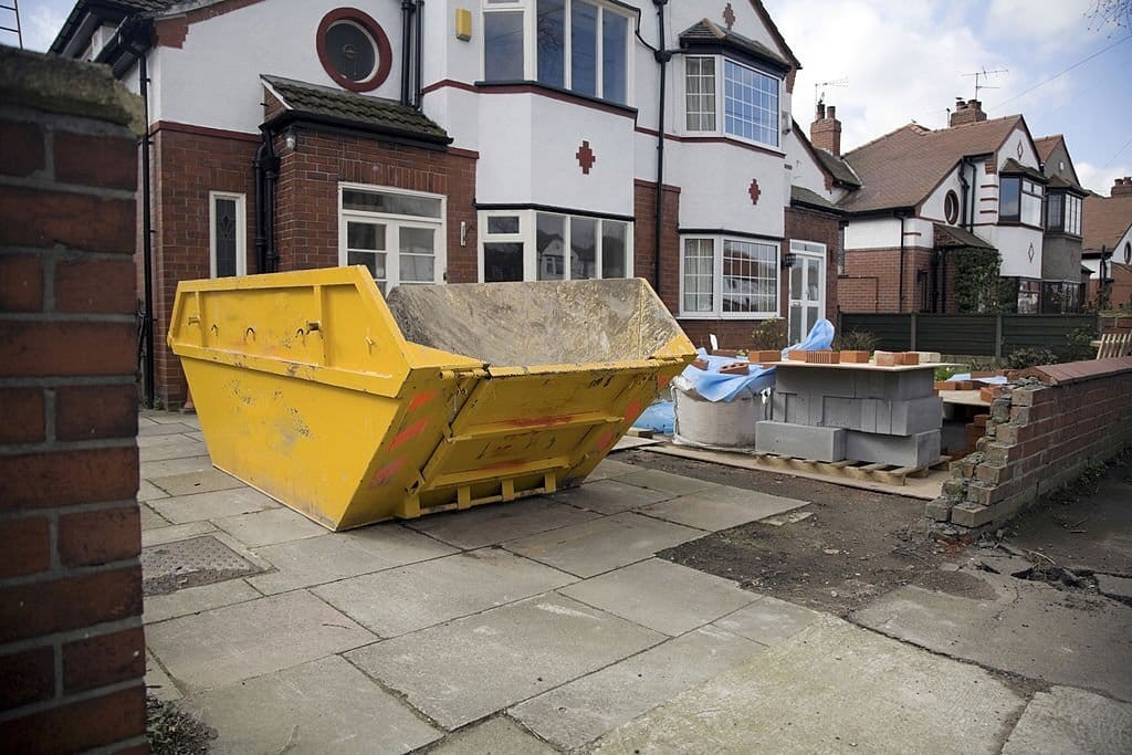 Home renovation in progress with a large yellow skip bin filled with debris in the driveway of a classic 1930s semi-detached house with red brick walls and white windows, signaling a house extension and property improvement project.