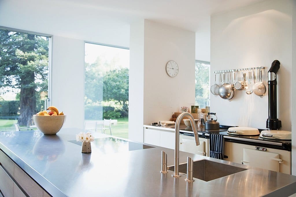 Sleek modern kitchen with stainless steel countertops, large floor-to-ceiling windows offering garden views, and hanging stainless steel pots and pans, exemplifying contemporary interior design.