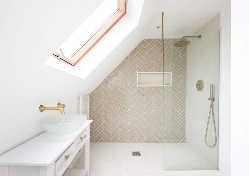 Minimalist bathroom design in a loft conversion featuring a herringbone patterned tile shower, a wall-mounted gold faucet, a vessel sink, and a skylight window for natural light.