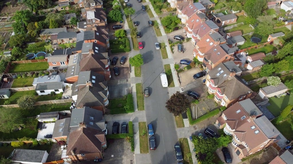 Aerial view of a tranquil suburban neighborhood showcasing a mix of 1930s semi-detached houses with driveways and modern extensions, interspersed with lush backyards and a variety of parked cars, reflecting typical residential community planning.