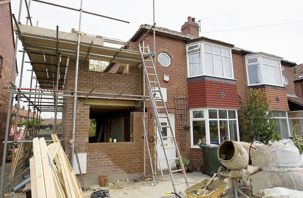 Active construction site at a red brick 1930s semi-detached house with scaffolding for an upper-level extension, featuring building materials, a ladder, and a cement mixer in the foreground, illustrating a suburban home double-storey side extension and renovation project.