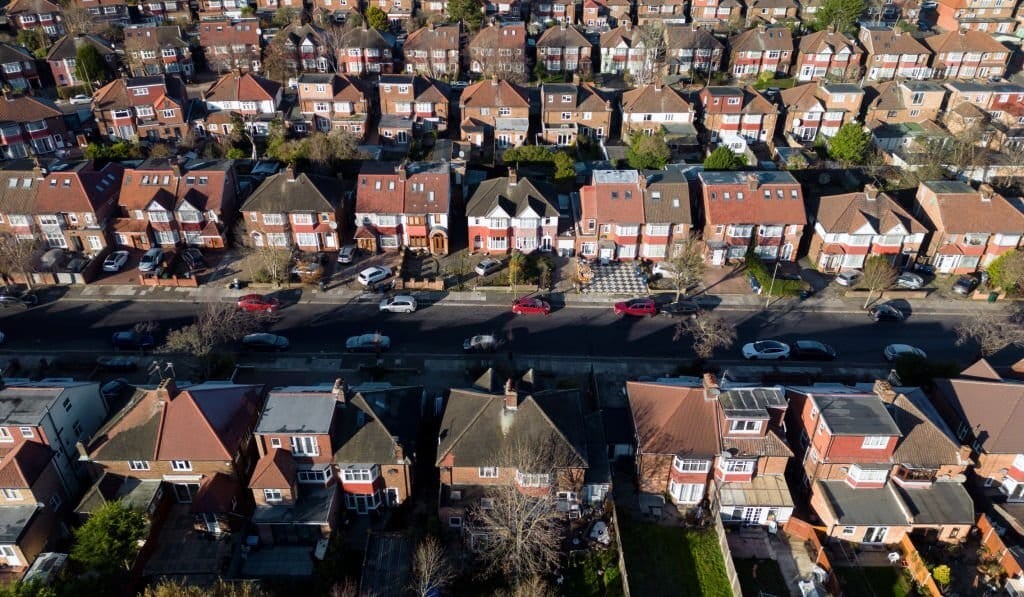Aerial shot of a densely packed suburban street in the UK with rows of 1930s red-brick semi-detached houses, featuring bay windows, tiled roofs, and extensions, interspersed with vehicles and tree-lined sidewalks.