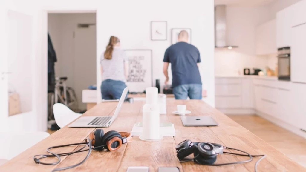 Two people in a shared kitchen and dining area, with laptops and headphones on a large wooden table.