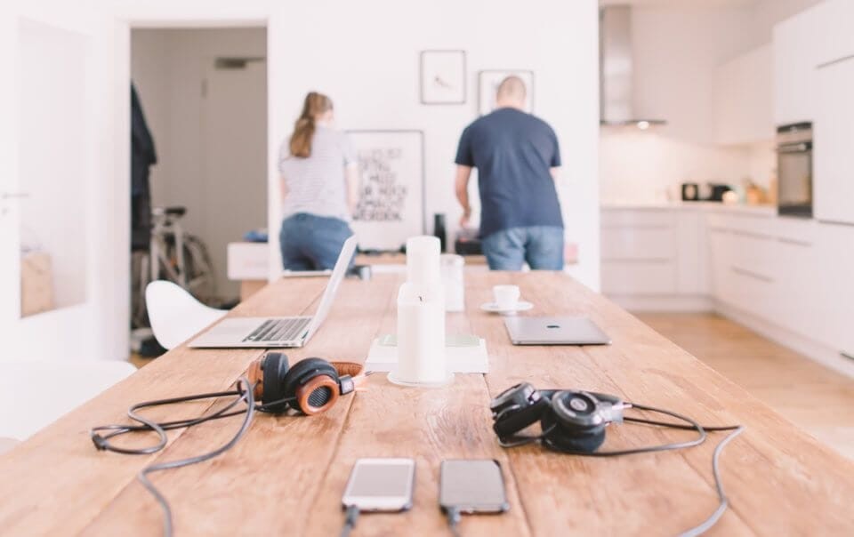 Two flatmates have stepped away from working at their dining table, leaving their laptops, phones and headsets lying about as they go about other tasks in their bright spacious apartment