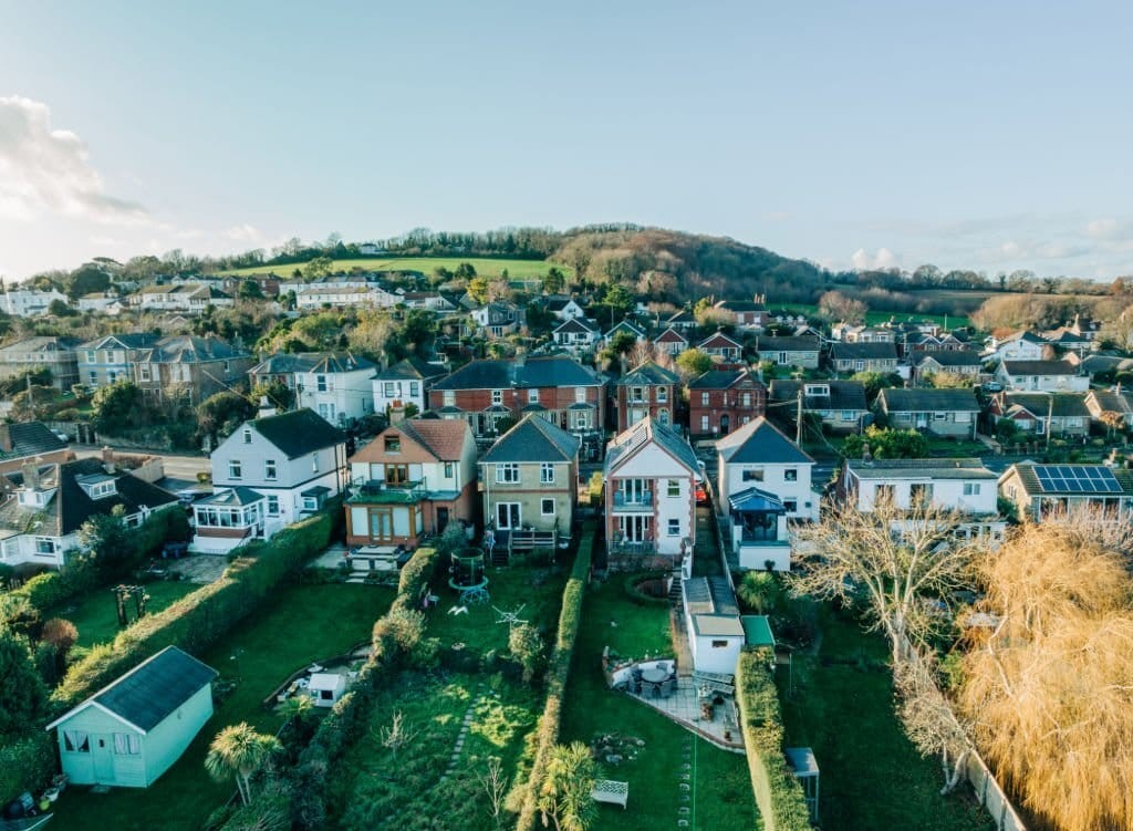 Aerial view of a serene residential area with a variety of detached and semi-detached homes, green lawns, and gardens, against a backdrop of rolling hills and clear skies.