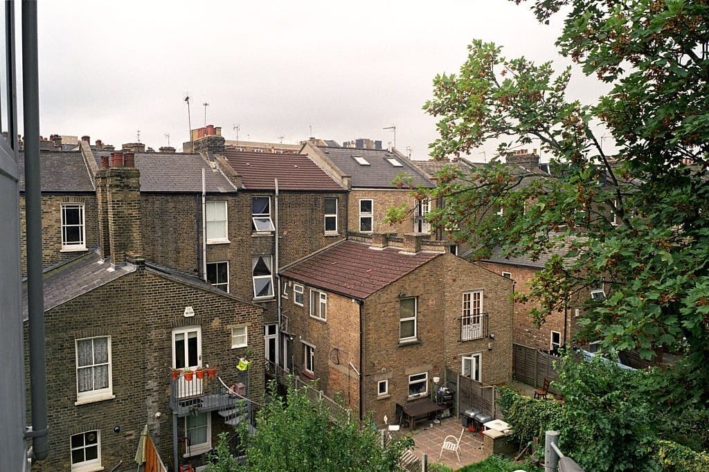 Overlooking a cluster of traditional London terraced houses with varying roof levels, surrounded by greenery on an overcast day, highlighting the dense urban living space.