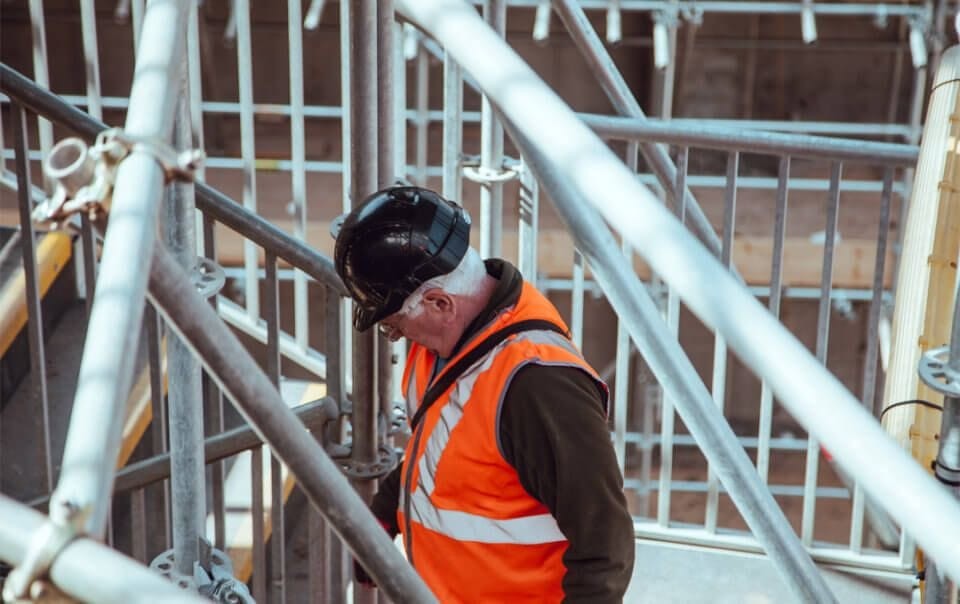 Contractor in an orange hi-vis vest and black hard hat walking up the stairs of a medium size building whilst the biggest scaffolding company in the UK finalise setting up