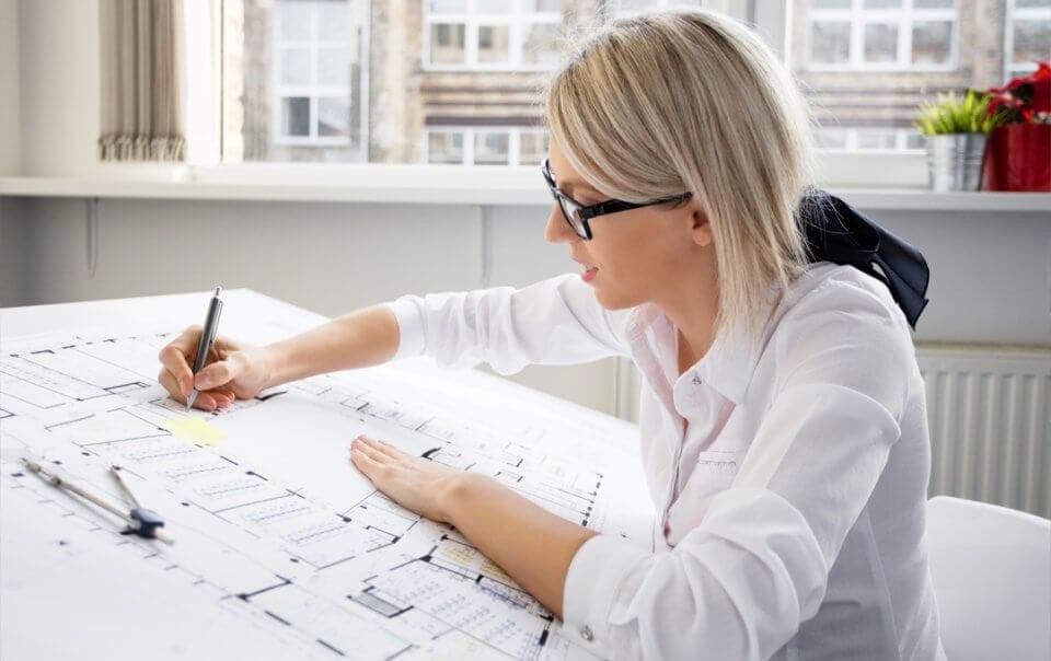 Female architect wearing a white blouse with her blonde hair tied in a ponytail with a black ribbon and wearing black rimmed glasses drawing with a pen on technical drawings printed in A3 on an architect drawing board