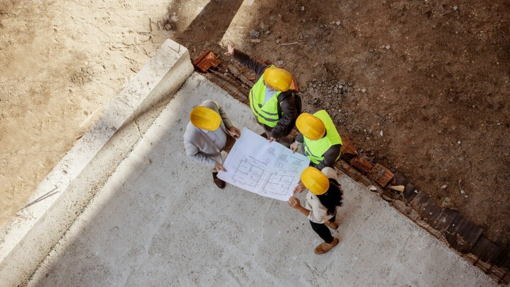 Aerial view of three construction workers with hard hats and high-visibility vests discussing over building plans on a construction site with exposed foundations.