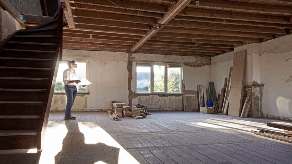 Professional architect reviewing blueprints in a spacious room under renovation with exposed wooden beams, natural light, and construction materials.