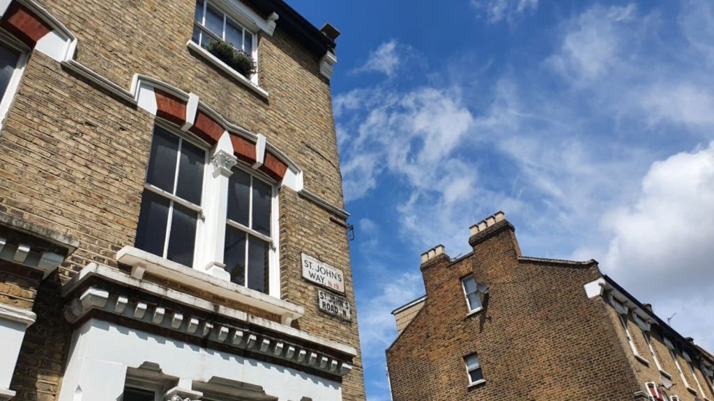 Victorian style brick corner buildings under a clear blue sky with white clouds, showcasing traditional London architecture with a street sign for St. John’s Way, Archway N19.