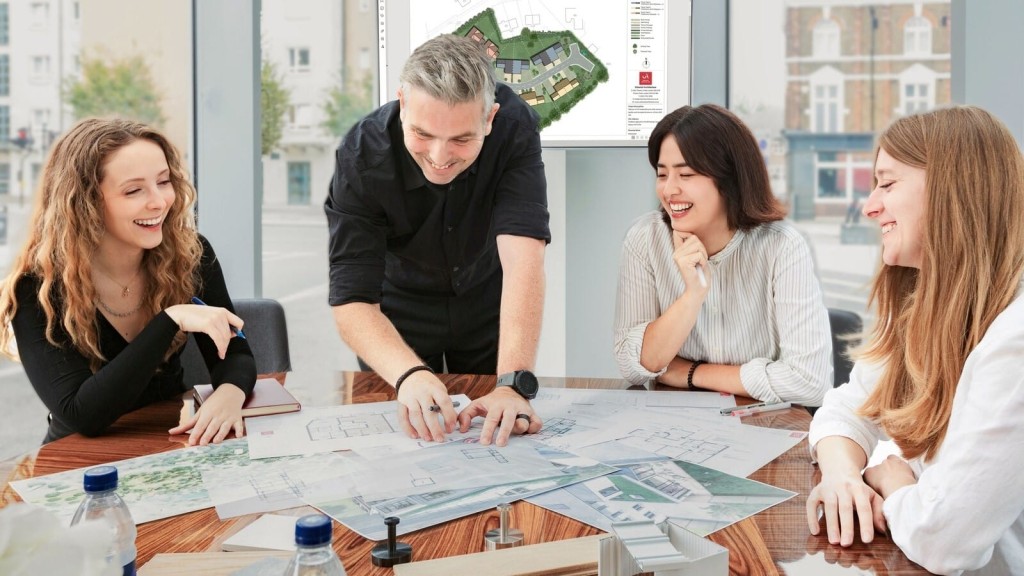 Group of four cheerful architectural designers in a well-lit office engaging in a lively discussion over architectural blueprints and a 3D building model, with urban streetscape visible through the window.