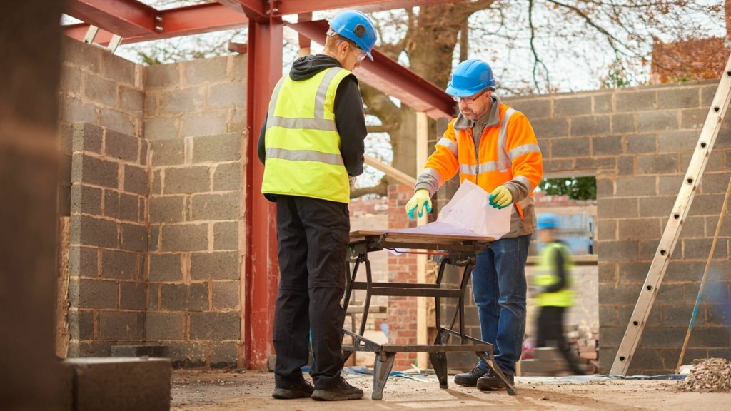 Two construction workers in safety gear examining architectural plans on a workbench at a building site, with brick walls and steel beams in the background.