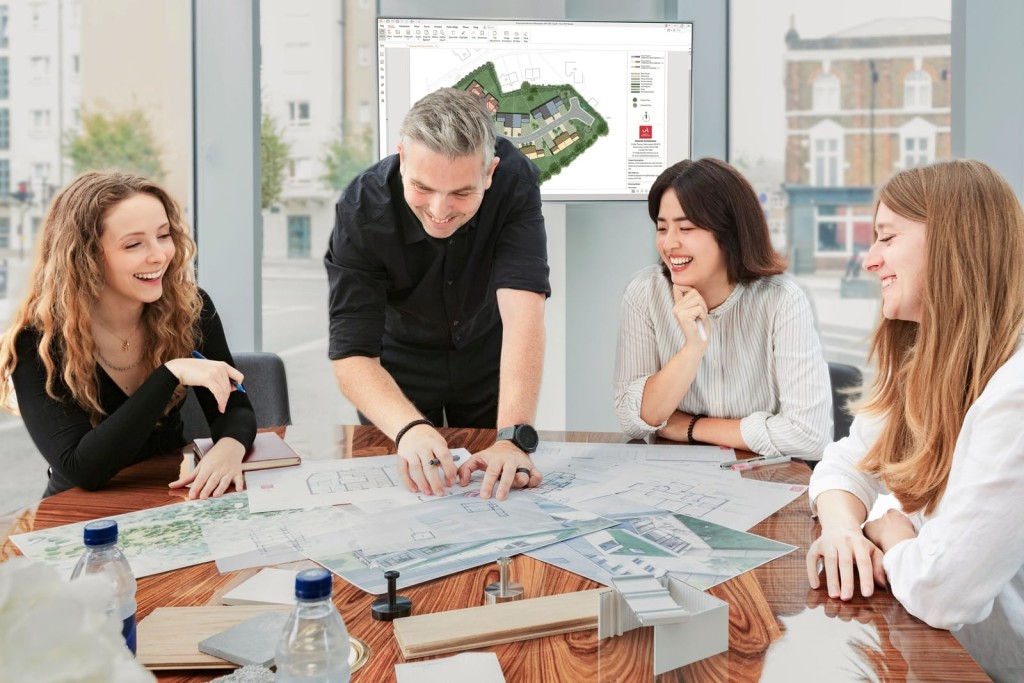 Team of architects smiling and collaborating over building blueprints on a table with a computer screen displaying 3D architectural software in the background, indicating a dynamic planning session.