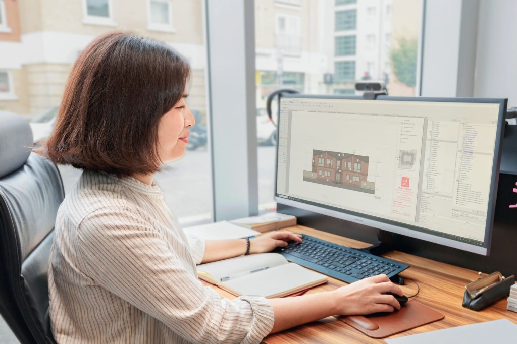 Professional female architect reviewing architectural 3D model on computer screen in a bright office setting, showcasing modern building design and planning.