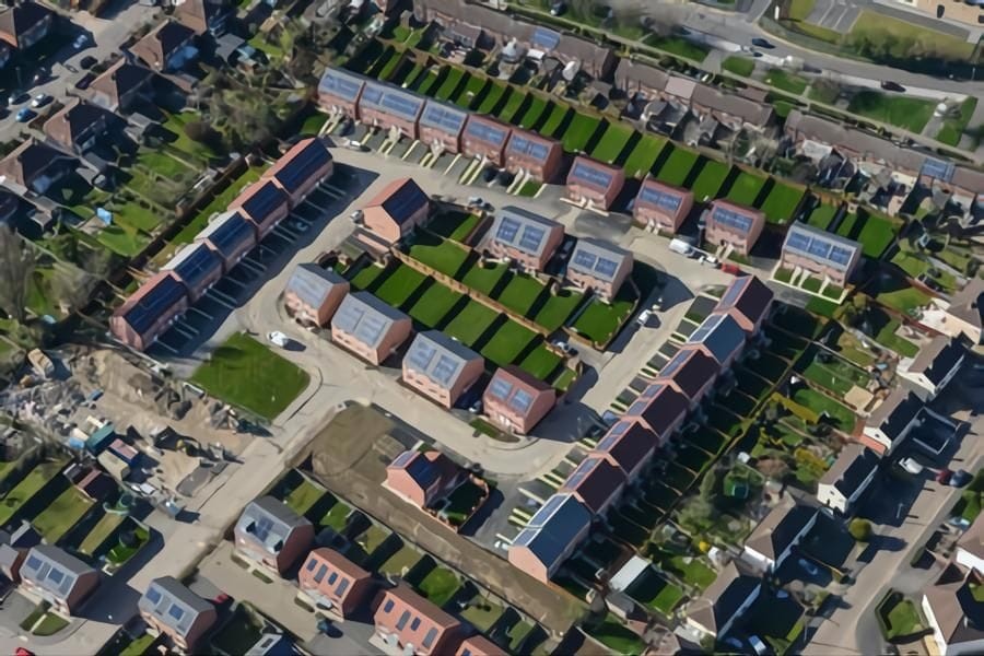 Aerial view of a suburban housing development showing rows of houses with solar panels on the roofs, neat gardens, and a clear road network, illustrating sustainable living in a residential community.