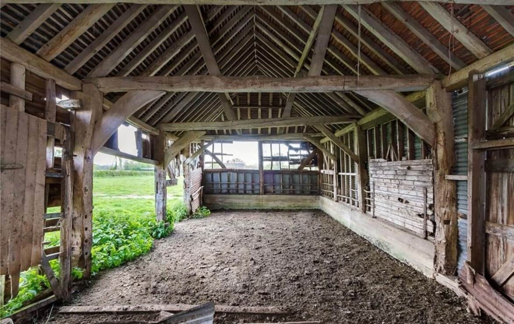 Interior of a rustic barn pre-renovation, showcasing exposed wooden beams and rafters, weathered wooden slats, and a view of the surrounding green fields, epitomising potential for rural barn conversions in the UK.