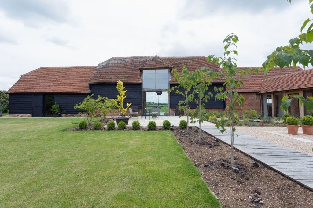 Modern barn conversion with large glass facade set in a landscaped garden, featuring a neatly manicured lawn, a stone pathway, and young trees, reflecting a blend of contemporary design and traditional country living.