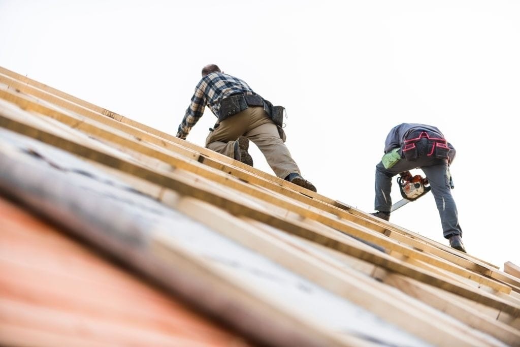 Construction workers in safety gear installing wooden roof beams on a barn conversion, highlighting skilled craftsmanship and renovation work in progress, set against a clear sky.
