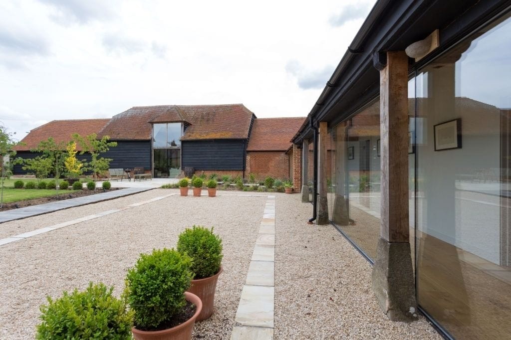 Modern barn conversion in the UK featuring a spacious gravel courtyard, traditional red brick walls, large glass windows, and a glazed walkway lined with potted boxwood shrubs, highlighting a blend of rustic architecture with contemporary design elements.