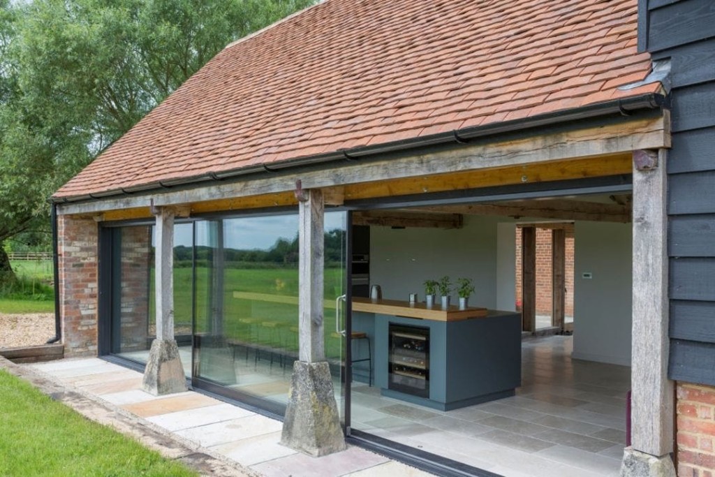 Renovated barn with an open-plan interior visible through full-height glass doors, featuring a modern kitchen island, against the backdrop of a serene UK countryside, exemplifying successful integration of contemporary design within traditional structures.