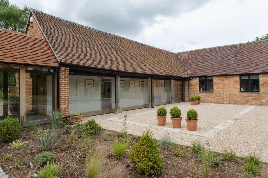 Elegant barn conversion in the UK with a spacious gravel patio, framed by large glass walkway and red brick walls, complemented by potted evergreen shrubs and a flourishing garden bed, illustrating a modern-rustic residential design.