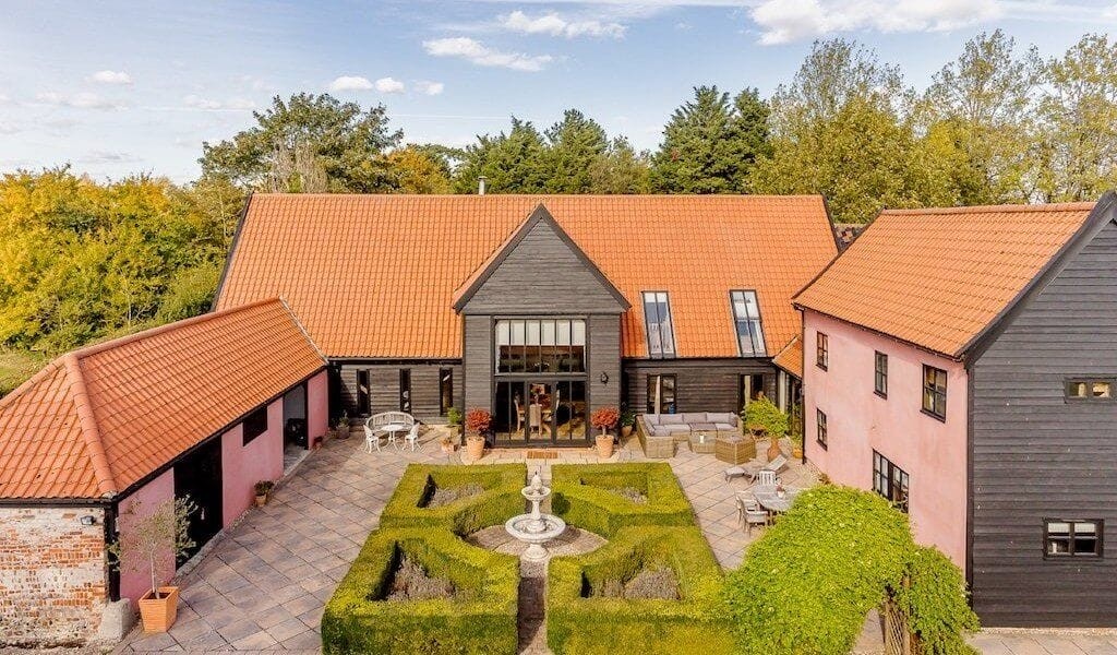 Aerial view of a luxurious barn conversion in the UK, featuring black and pink facades with terracotta roofs, a manicured garden with a central fountain, and outdoor seating areas, nestled in a tranquil woodland setting, perfect for upscale rural retreats.
