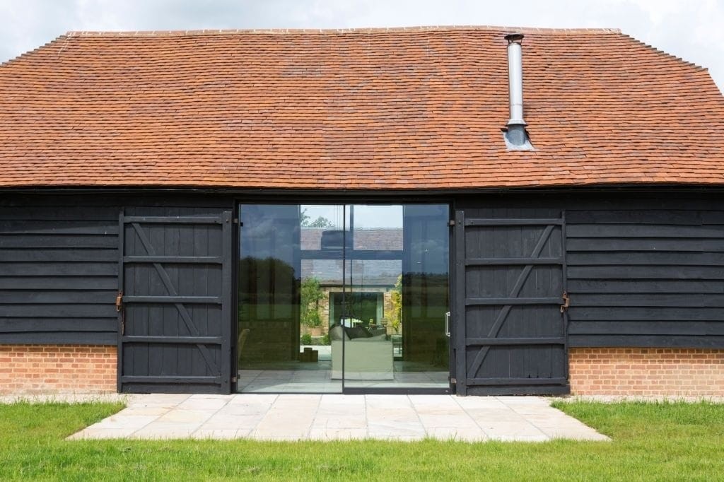 Elegant barn conversion with black timber cladding and a terracotta tile roof, featuring large glass doors flanked by original wooden barn doors, set in a lush green landscape, reflecting a fusion of classic rural architecture with modern design in the UK.