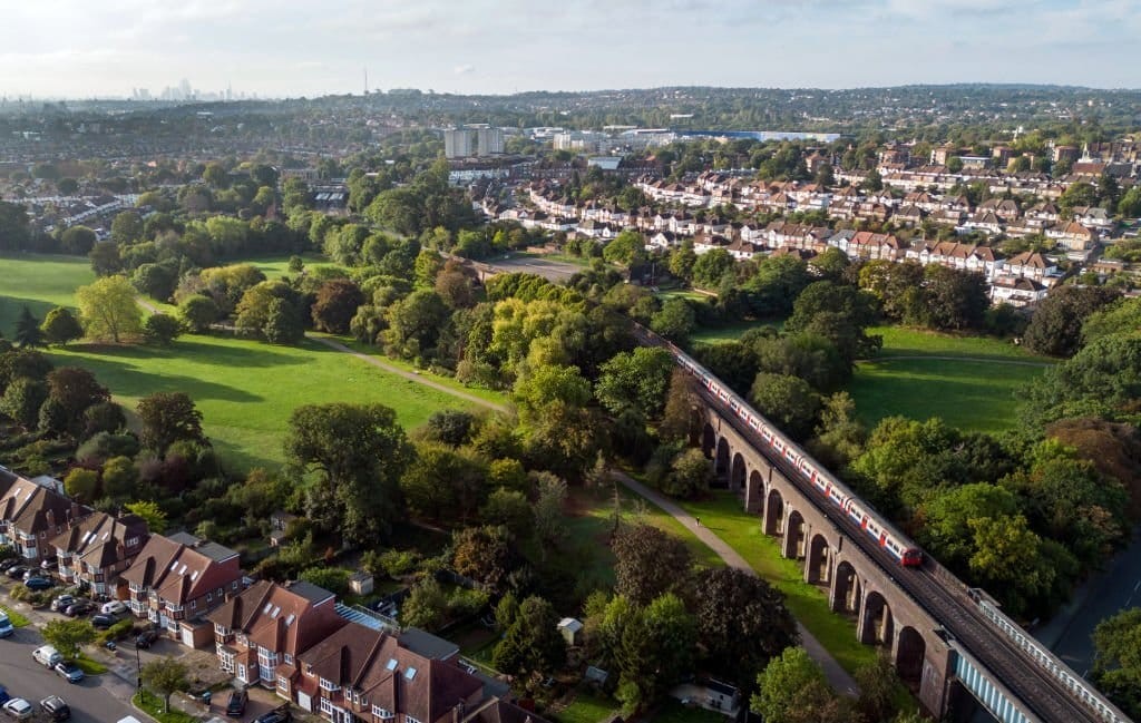 Aerial view of a UK suburban area with residential houses, a green park, and a red train crossing a viaduct, with a city skyline in the distance.