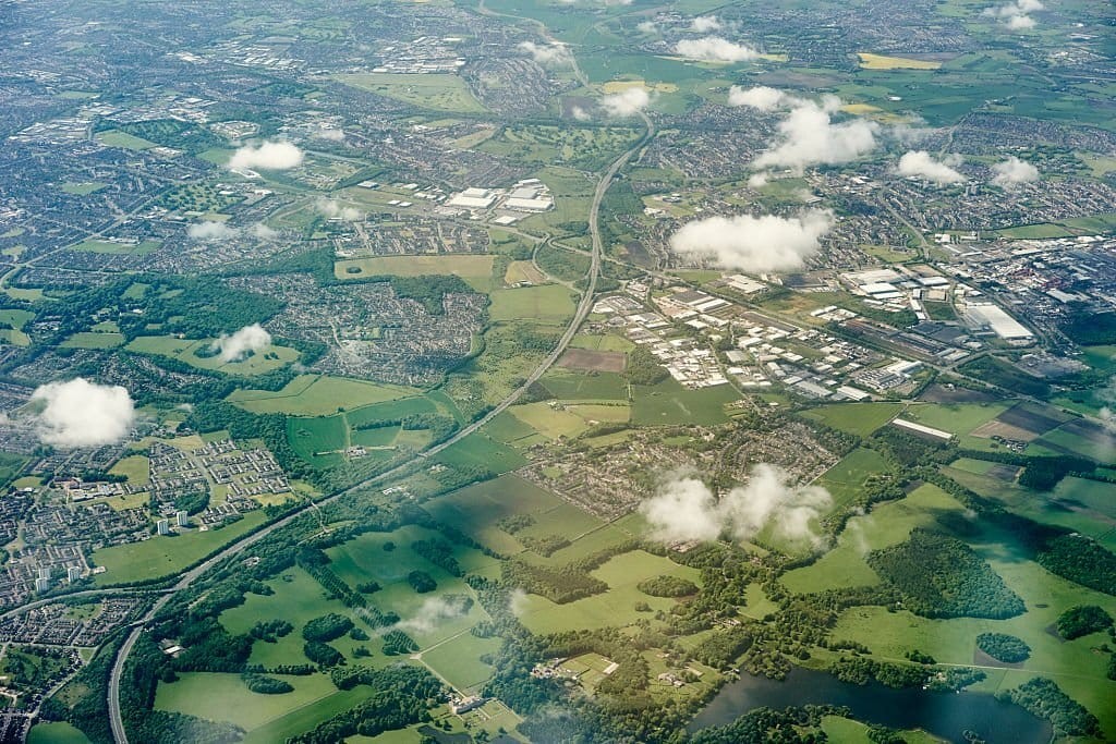 Aerial view of the English countryside with patches of green fields, clusters of houses, industrial buildings, and fluffy white clouds casting shadows on the landscape.