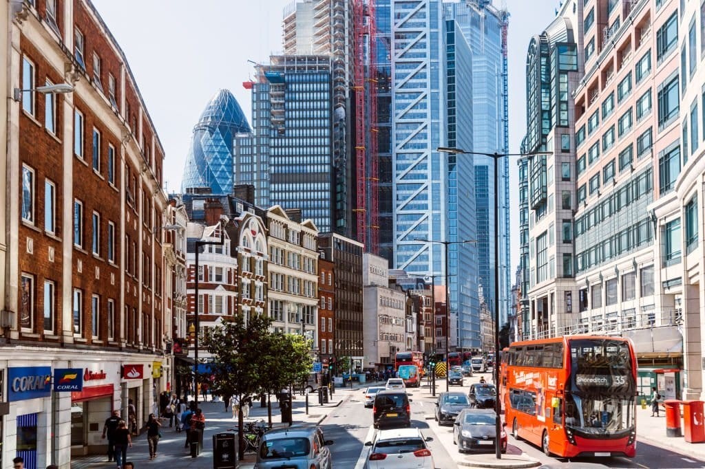 Bustling London street scene with diverse architecture, showcasing the contrast between historic buildings and modern skyscrapers under a clear blue sky, with busy traffic and a bright red double-decker bus en route to Shoreditch, embodying the dynamic urban life.