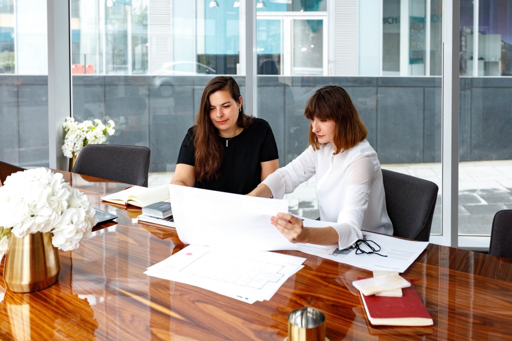 Two professional interior designers reviewing architectural plans at a modern office with large windows. The workspace features a polished wooden table adorned with design books and fresh white flowers, reflecting a blend of creativity and professionalism.