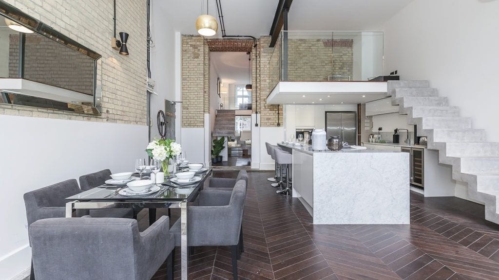 Modern open-plan kitchen and dining area. The space features a sleek white kitchen island with marble countertops, contemporary grey dining chairs around a glass table, and a mezzanine level. Exposed brick walls and a mix of industrial and minimalist design elements highlight the expertise of top London interior designers in creating chic, functional living spaces.