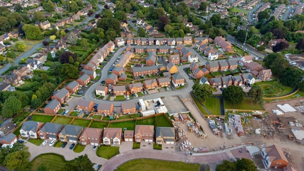 Aerial perspective of a suburban housing development with a variety of detached and semi-detached homes, featuring construction areas with materials and machinery for ongoing expansion, amidst landscaped greenery.