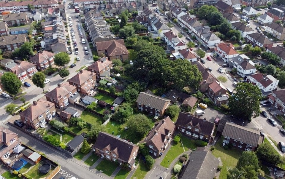 Aerial view of a residential neighborhood showing a pattern of detached and semi-detached houses with red-tiled roofs, interspersed with green lawns and trees, depicting typical suburban planning and development.