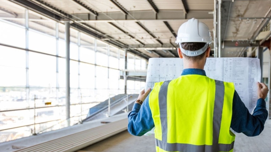 Construction engineer in a high-visibility vest and safety helmet reviewing architectural blueprints at a construction site with structural steelwork in the background, symbolising active project management.