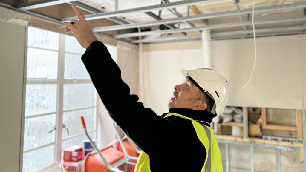 A construction worker in a hard hat and high-visibility vest inspects the interior of a building, pointing at the ceiling structure as part of building control checks.