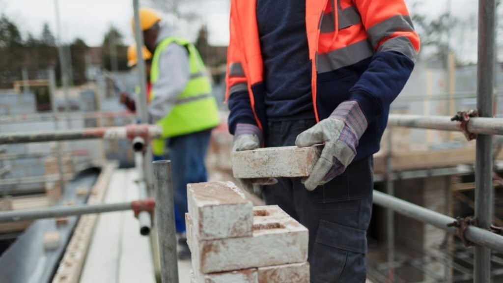 A construction worker holds a brick on a scaffold, with others in high-visibility vests working in the background on building control.