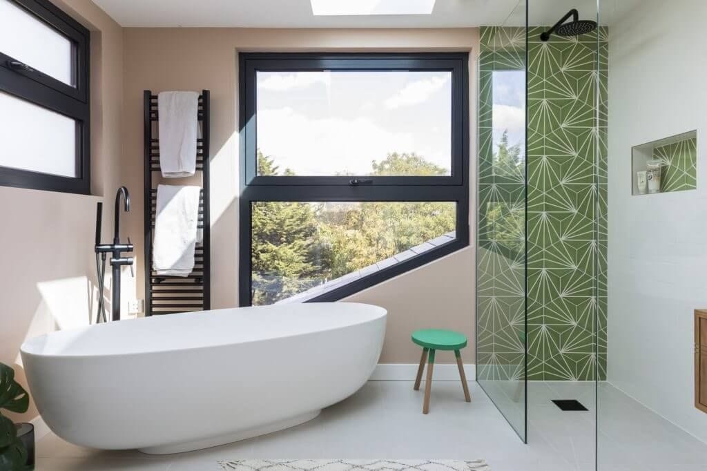 Modern bathroom in a dormer extension with a freestanding white bathtub, black fixtures, and a heated towel rail. The room features large windows providing ample natural light, green geometric tiles in the shower area, and a sleek glass shower enclosure. A small green stool adds a pop of color to the stylish, minimalist design.