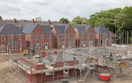 New housing development under construction in the UK, showcasing partially built brick houses with scaffolding and construction materials on-site, surrounded by mature trees and a residential neighbourhood.