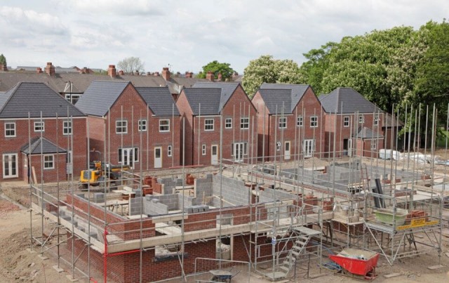 Image cover for the article: New housing development under construction in the UK, showcasing partially built brick houses with scaffolding and construction materials on-site, surrounded by mature trees and a residential neighbourhood.