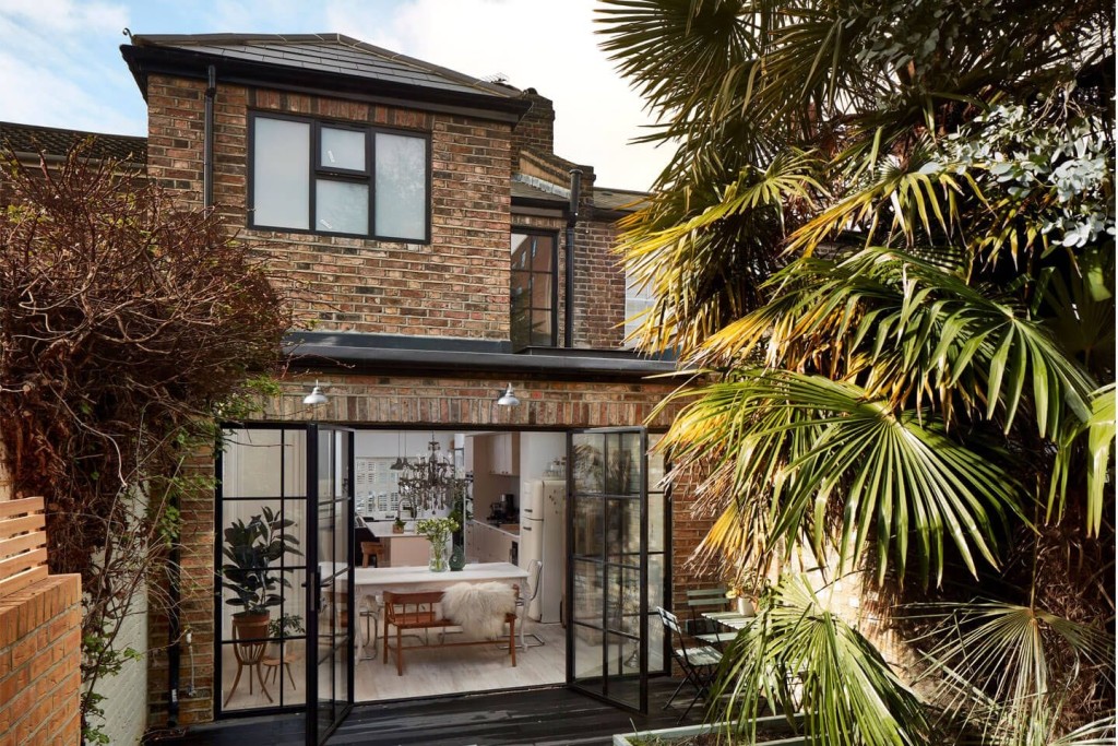 Rear view of a two-storey brick house showcasing a stylish home extension with large black-framed bifold doors leading to a cosy, modern kitchen and dining area. The lush garden features a variety of green plants and palm trees, enhancing the inviting outdoor space.