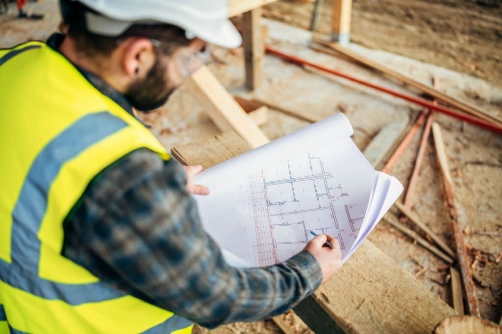 Contract adminstrator reviewing the technical drawings from the lead architects whilst in the middle of the construction site with loose wood chippings, wood planks and tubes but remaining safe in a high-vis jacket, protective glasses and a white hard hat