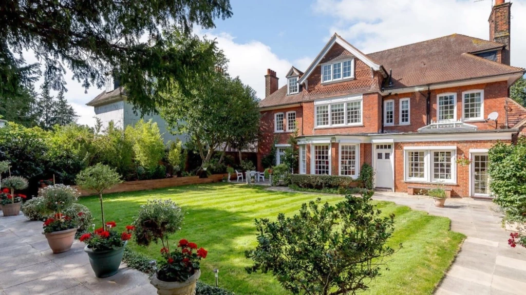 A grand red-brick Grade 2 listed building set within the Green Belt, featuring traditional sash windows, a gabled roof, and a beautifully maintained garden with a large lawn, mature trees, and potted plants lining the stone patio.