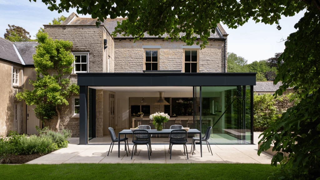A beautifully restored Grade 2 listed building with a striking modern rear extension featuring floor-to-ceiling glass walls and a black-framed flat roof, seamlessly connecting the contemporary kitchen–dining space to a landscaped garden with a sleek outdoor dining area.