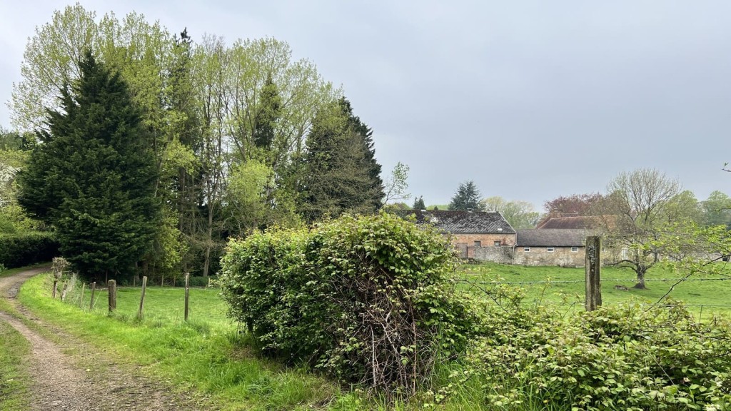 A scenic rural landscape featuring a dirt path alongside lush greenery and a fence leading to a brick building in the background. This image represents land that could be promoted for development through the call for sites process. It reflects the potential of agricultural or semi-rural areas being considered for housing or other land uses under updated planning policies. Perfect for illustrating the steps that occur after a site is selected for development, including feasibility studies and planning evaluations as part of the local plan preparation.