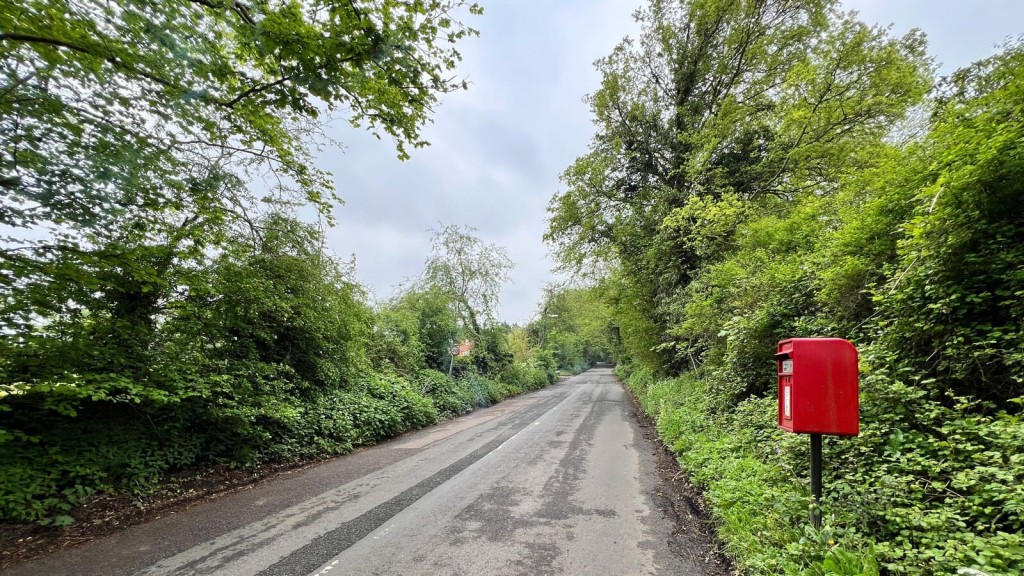 A quiet rural road bordered by lush greenery and trees, with a classic red postbox in the foreground. This image highlights the potential of greenfield and rural sites for development, fitting into the context of the "call for sites" process, where landowners and developers can promote their land for inclusion in local housing plans. Ideal for showcasing countryside locations that may be considered for sustainable housing projects under updated planning policies.